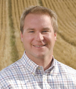 A headshot of Agronomist and farmer Steve Larocque as he stands before a large field with neat yellow rows.