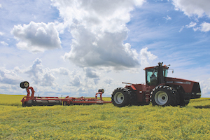 A roller crimping tractor is pictured crimping yellow blossom sweet clover, a yellow plant in a large field. The sky is blue with fluffy clouds rolling by.