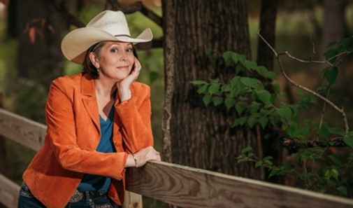 A Caucasian woman leans on a fence near a forest. She is wearing a tan cowboy hat and a vibrant orange blazer. 