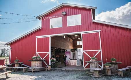 A large red and white barn with doors open to show the wedding venue inside. Barrels are outside decorated with flowers. 