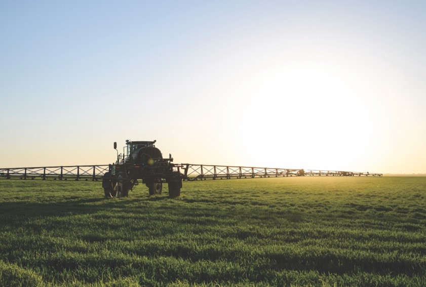 A large tractor with a wide sprayer drives along a flat field that sprawls into the horizon. The sun is setting in the distance. 