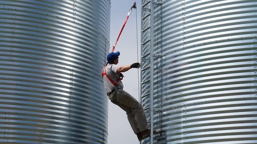 Farmer climbs a ladder up the side of a grain bin, clipped onto a safety line with a harness.