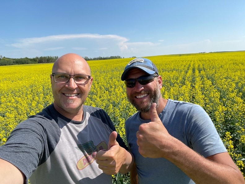 Two men stand in front of a thriving bright yellow canola field. They are both smiling and giving the thumbs up sign.