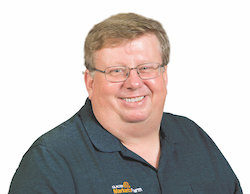 A portrait of Bruce Bernett, who is pictures wearing a navy polo shirt and glasses, smiling brightly. He is the Director Of Markets And Weather Information for Glacier Farmmedia.